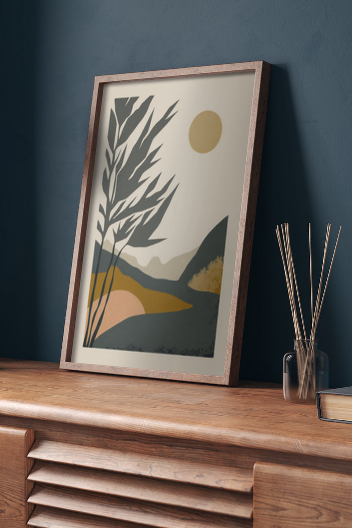 Modern abstract poster featuring stylized hills, sun and foliage in earthy tones, displayed in a wooden frame on a sideboard
