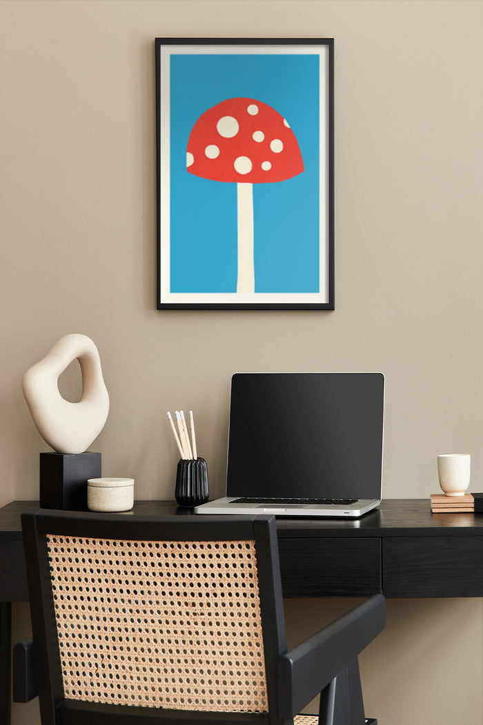 Minimalist red mushroom with white dots poster framed on wall above a workspace with laptop, modern sculpture, and office supplies